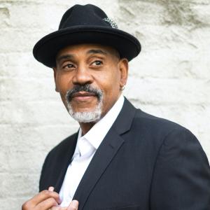 Allan Harris in a hat and jacket and white shirt, looking off to the side with a salt and pepper beard. He stand in front of a white-painted brick wall.