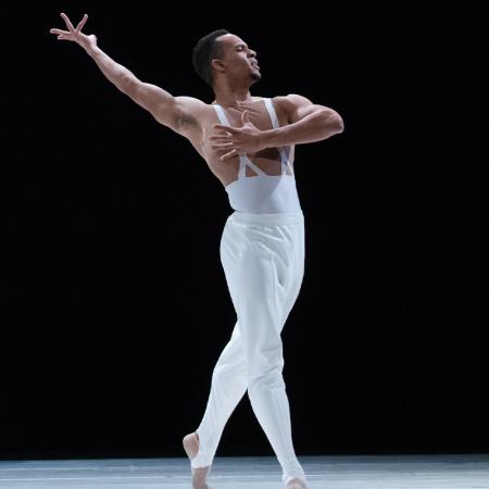 A member of Ballet Hispanico in white, one arm held up in the air, the other across his chest in a dramatic pose