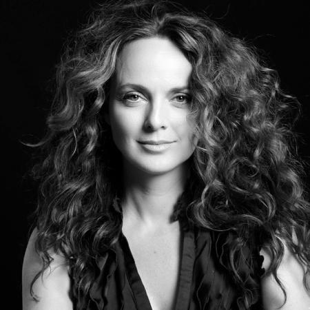 Black-and-white portrait of Melissa Errico, with voluminous curly hair, wearing a sleeveless textured top, looking confidently into the camera against a dark background.